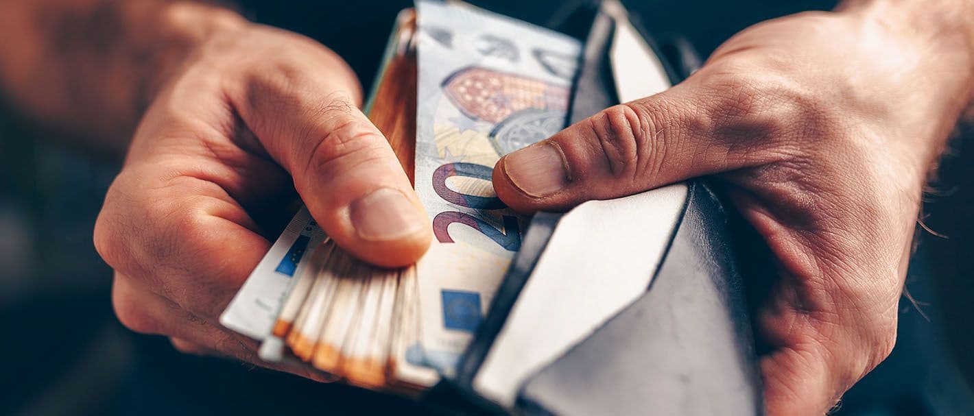Nahaufnahme der Hände eines Mannes, die einen geöffneten Geldbeutel halten und einen aufgefächerten Stapel Geldscheinen, der zur Hälfte darin steckt.