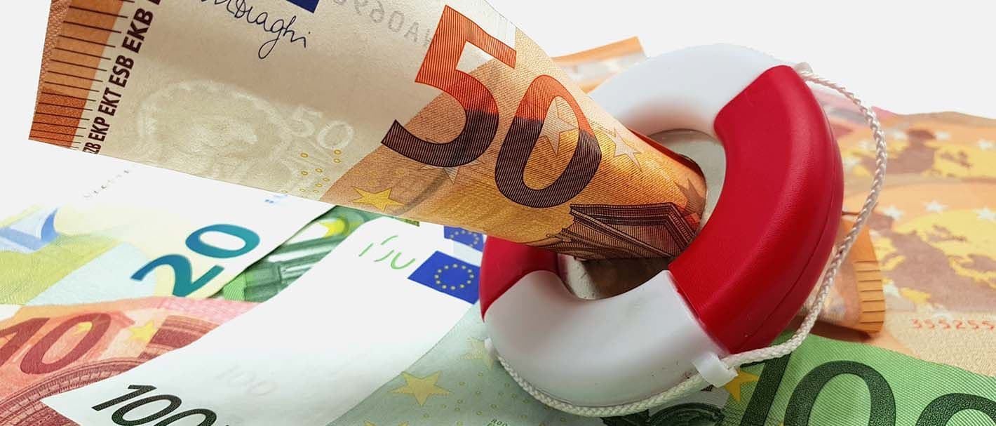 Ein 50-Euroschein steckt in einem Rettungsring. das Arrangement liegt auf einer Fläche, die mit 100-, 50- 20- und 10-Euro-Scheinen bedeckt ist.