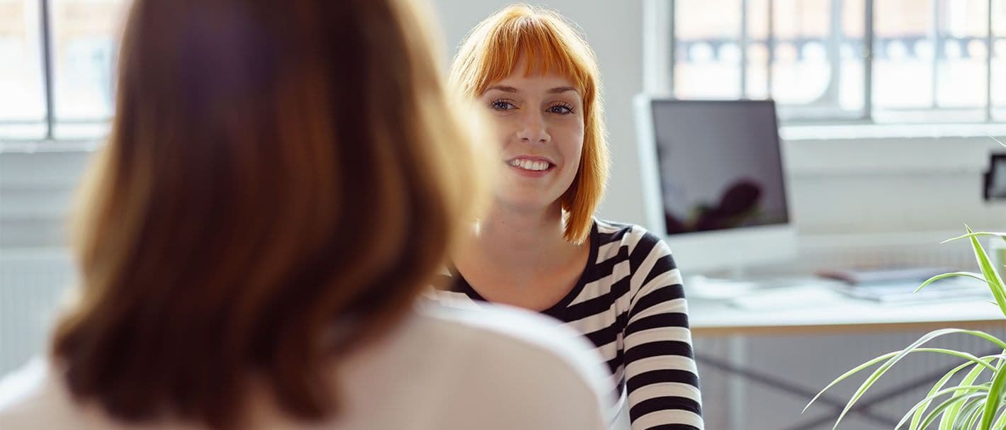 Zwei Frauen sitzen sich in einem Büro bei einem Gespräch gegenüber. Die eine Frau ist dem Betrachter zugewandt, die andere ist von hinten abgebildet.